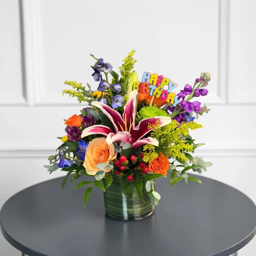 Colorful birthday flower arrangement with lilies, delphinium, roses, and a Happy  Birthday pick.